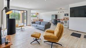 A modern living room with a sofa, armchair, and ottoman, a dining area, and a kitchen with a large window.