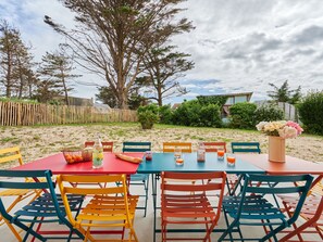 Un patio con una mesa dispuesta para dos personas, rodeado de sillas coloridas, y vista a una zona arenosa y árboles al fondo.