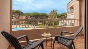 Un balcon avec deux fauteuils, une petite table avec une bouteille et des verres, et une vue sur une piscine et des immeubles.