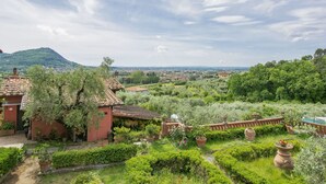 A lush garden with terracotta pots, a red building, and a mountainous backdrop. - Beautiful apartment for 2 guests with hot tub, pool, A/C, WIFI, TV, patio and pets allowed (Montecatini Terme)