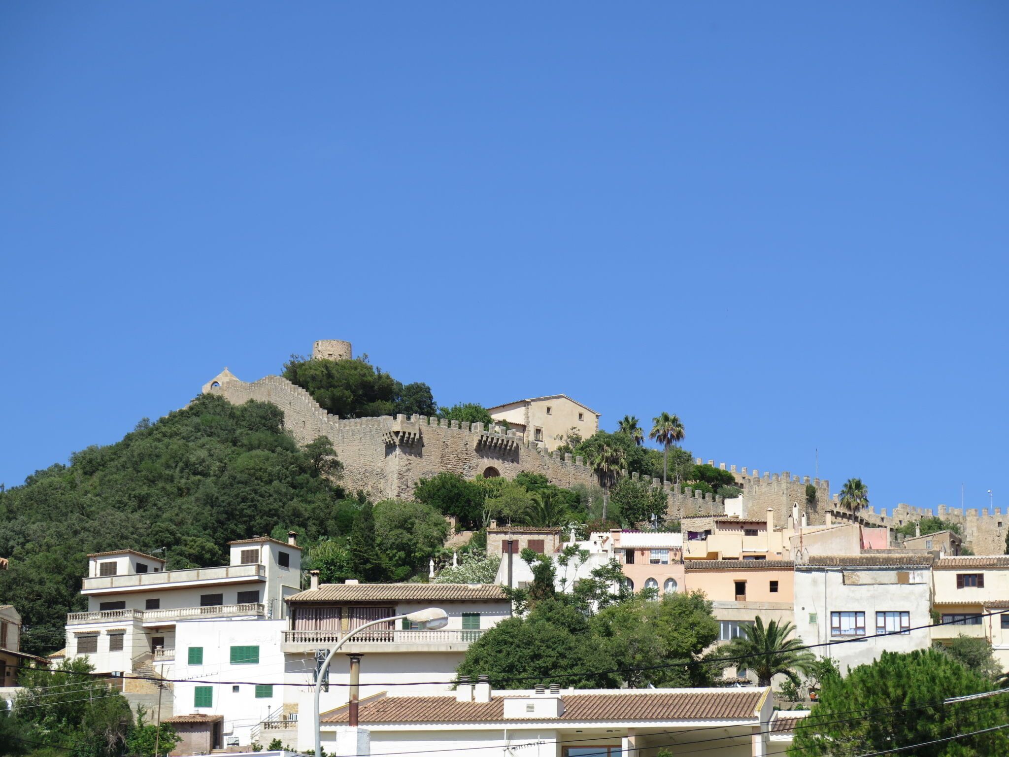 A historic hilltop castle with a fortified wall, surrounded by residential buildings with traditional architecture.