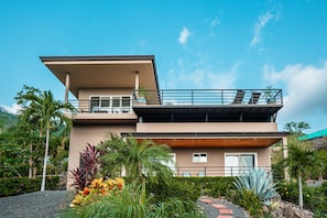 A modern two-story house with a balcony, surrounded by tropical plants and a clear sky.