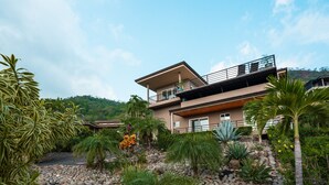 Ein modernes, zweistöckiges Haus mit Balkon, umgeben von einem Garten mit verschiedenen Pflanzen und steinigem Untergrund.