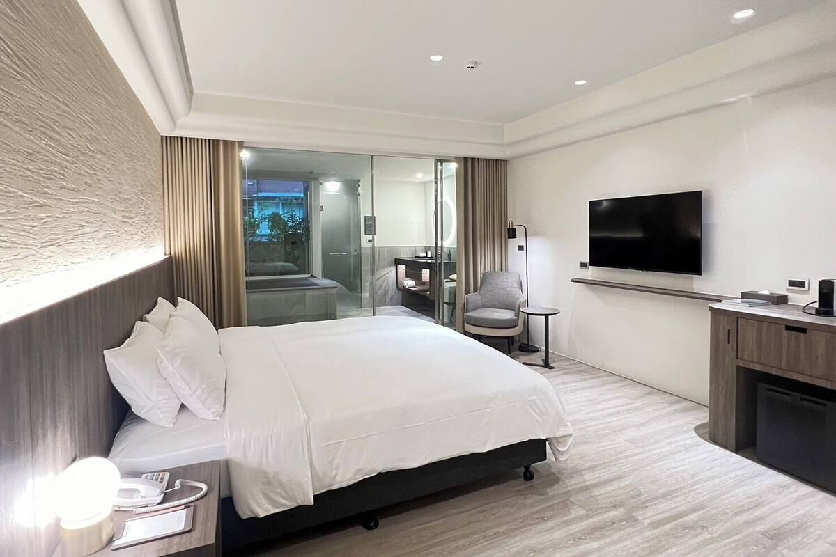 A modern hotel room with a large bed, a TV, a desk, and a glass shower area.