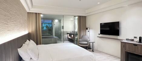 A modern hotel room with a large bed, a TV, a desk, and a glass shower area.