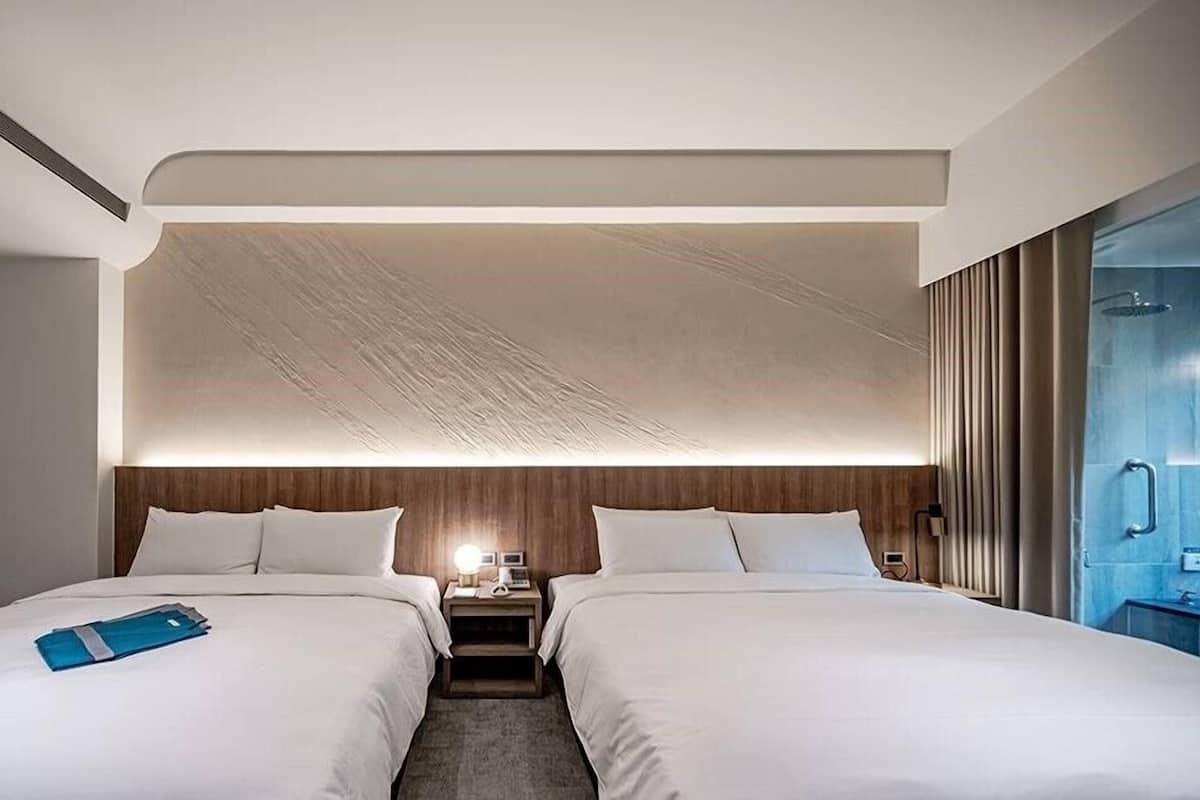A hotel room with two beds, a wooden headboard, a nightstand with a lamp, and a bathroom with a shower and mirror.