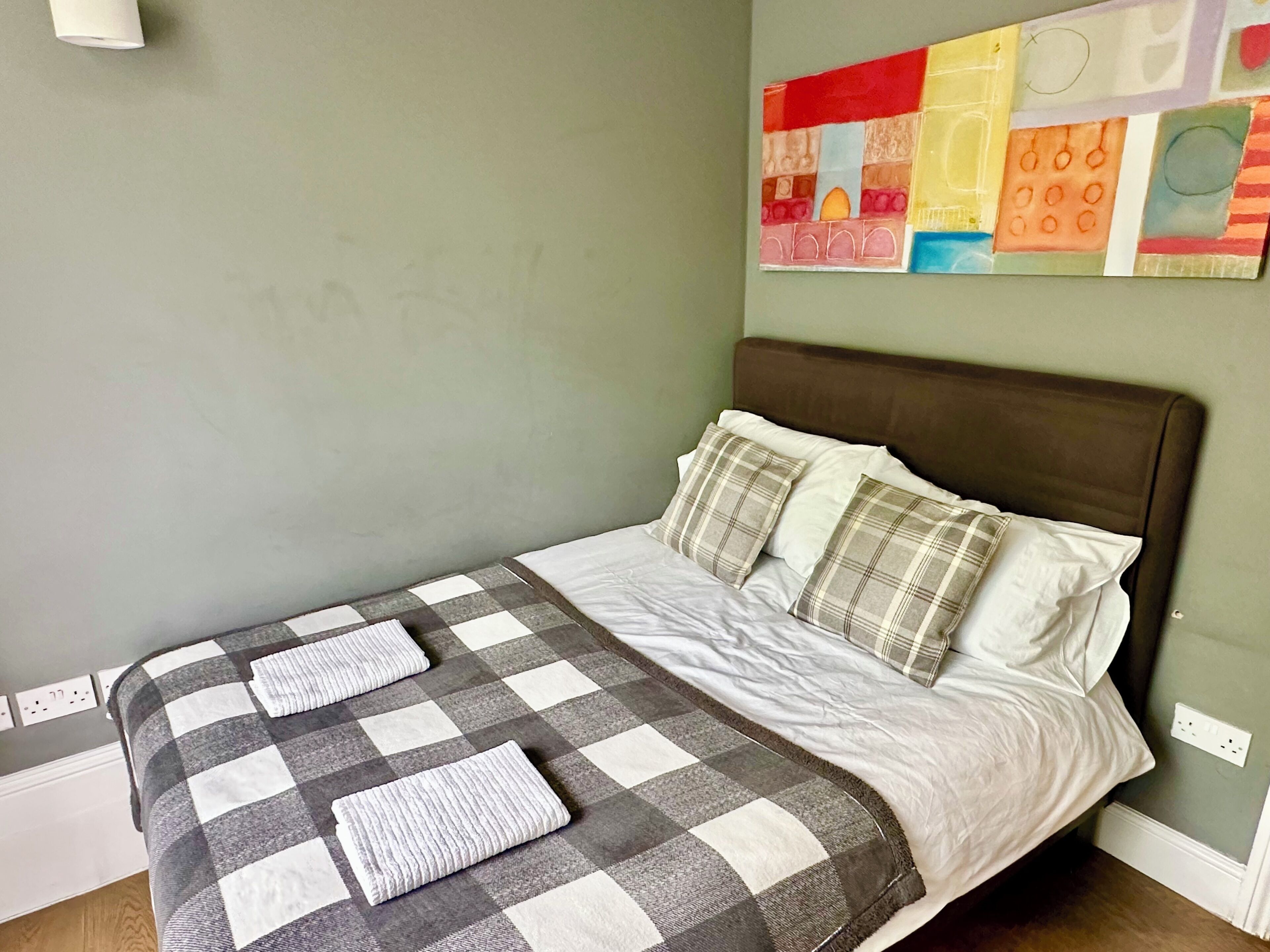 A neatly made bed with a checkered blanket and pillows, a brown headboard, and a colorful abstract painting on the wall.