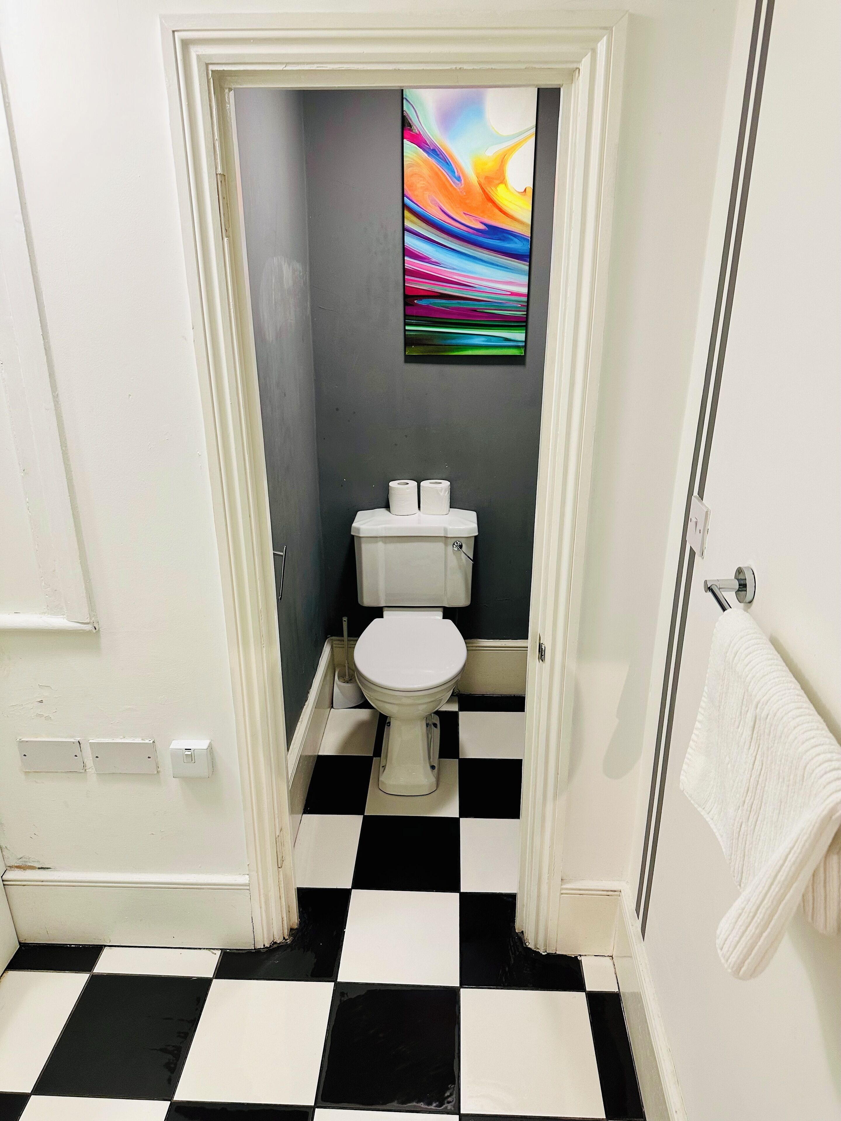 Um banheiro com piso xadrez preto e branco, vaso sanitário branco e um quadro abstrato colorido na parede.