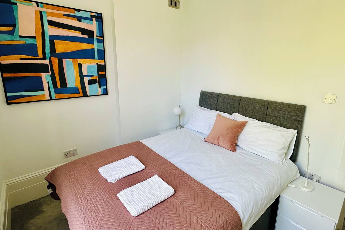 A neatly made bed with a brown quilt, white pillows, and a grey headboard. A white bedside table with a lamp is to the right.