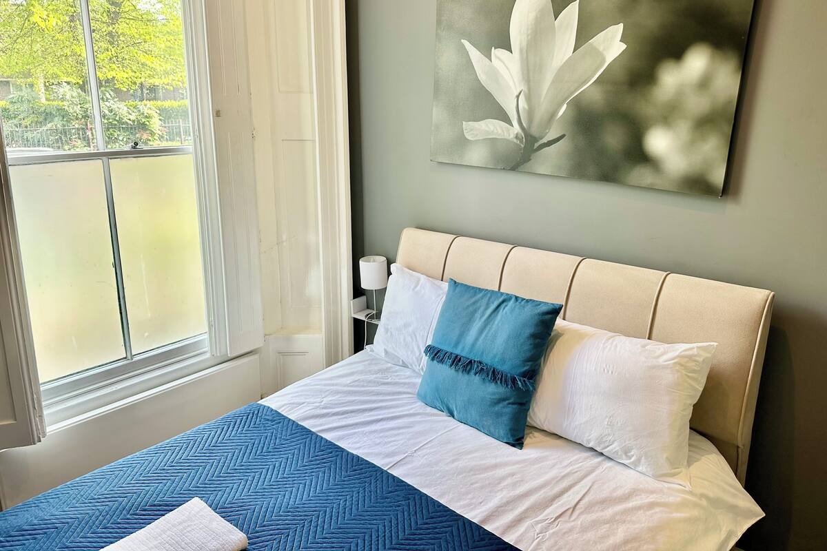 A neatly made bed with a blue and white bedspread, a single blue pillow, and a white pillow. A painting of a flower hangs on the wall above the bed.