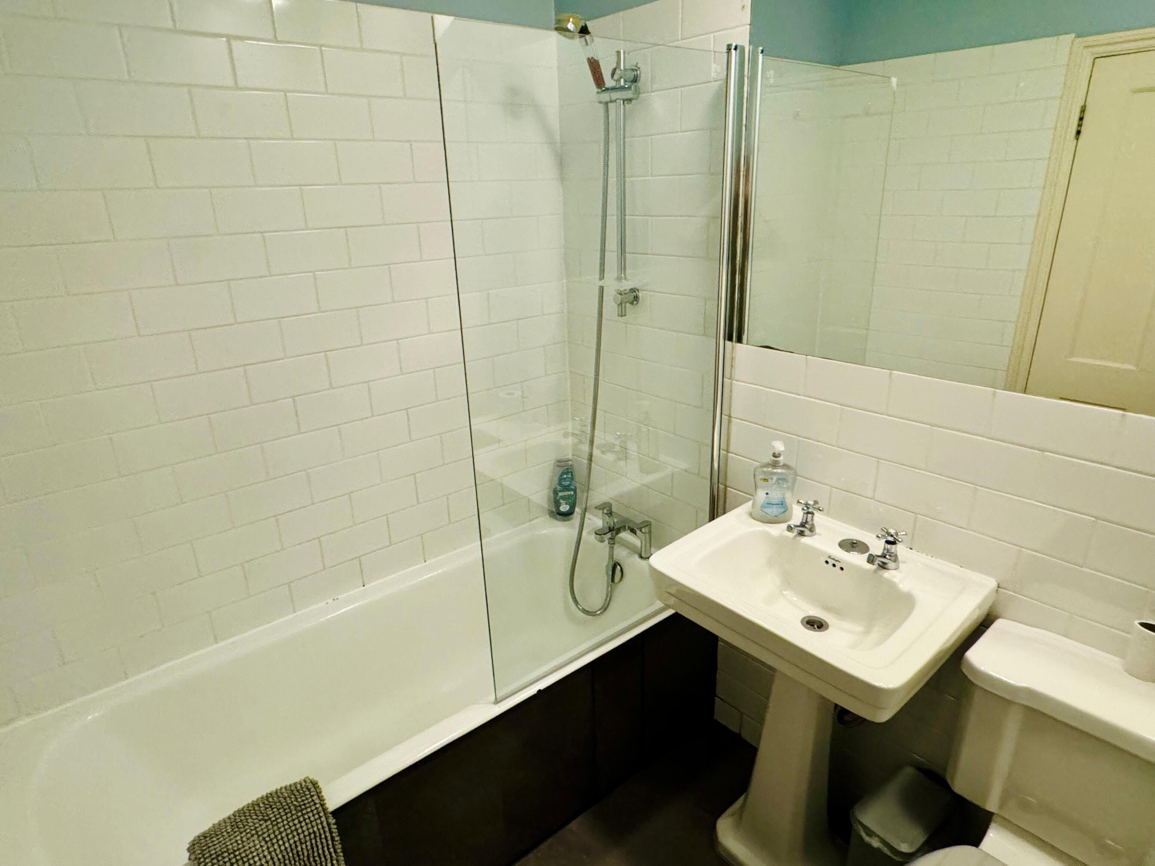 A bathroom with white tiled walls, a glass shower enclosure, a pedestal sink, and a toilet.