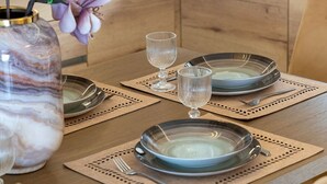 Una mesa de comedor dispuesta con platos, vasos y cubiertos, con un florero con flores a la izquierda.