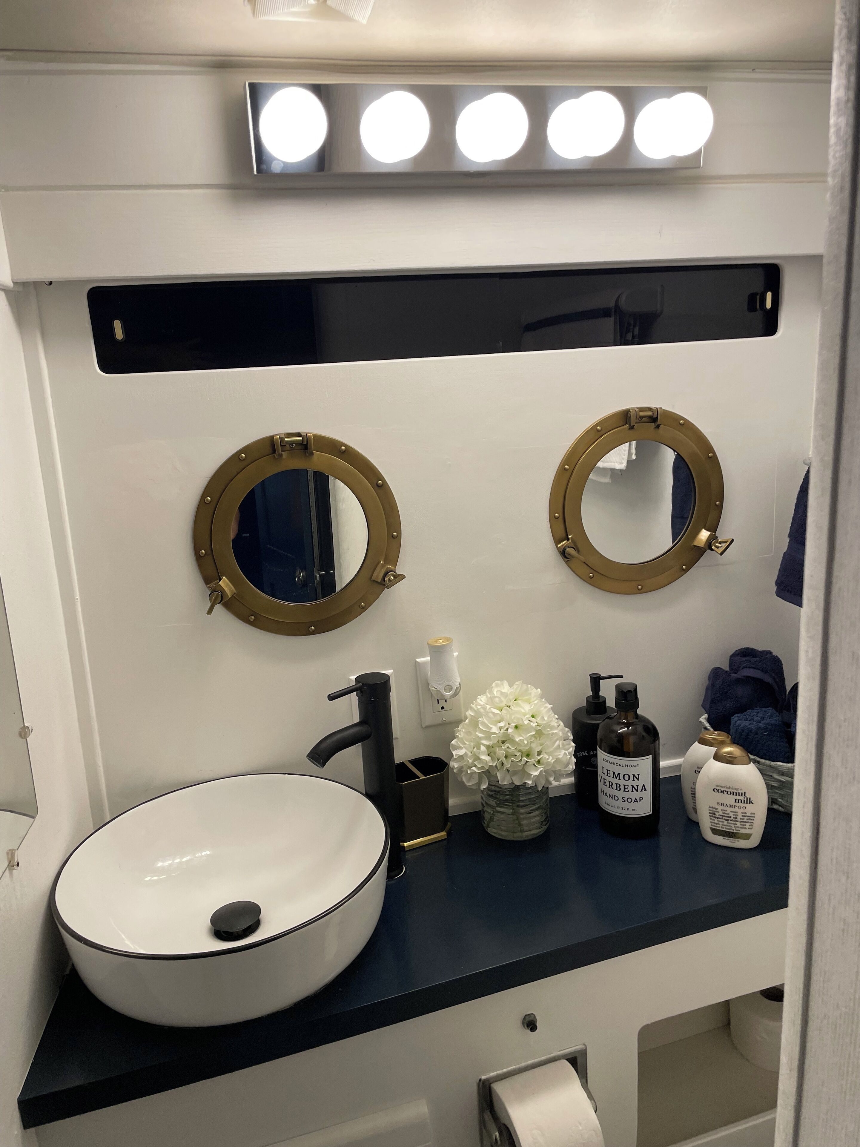 Une salle de bain avec un lavabo blanc, deux miroirs ronds à cadre doré, un robinet noir et un plan de toilette bleu. On y trouve des articles de toilette, notamment un flacon de shampoing et un distributeur de savon.