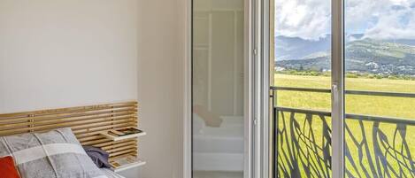 A bedroom with a bed, a nightstand, and a sliding glass door leading to a balcony with a view of the countryside.