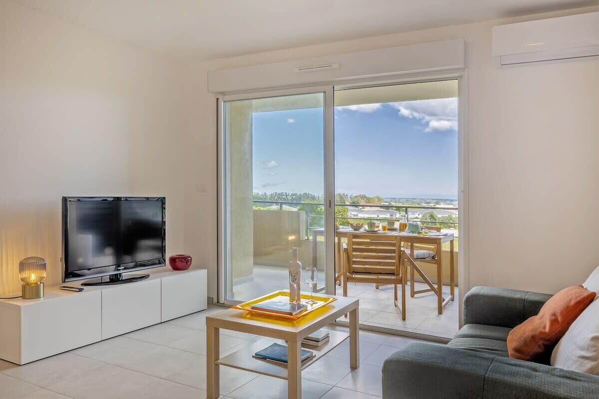 Ein modernes Wohnzimmer mit einem Flachbildfernseher auf einer weißen Konsole, einem Glastisch und einem grauen Sofa. Ein Balkon mit Essbereich ist ebenfalls vorhanden.