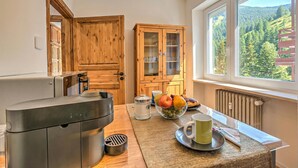 Een keuken met een koffieapparaat, houten kastjes, een fruitschaal en uitzicht op een bos door een raam.