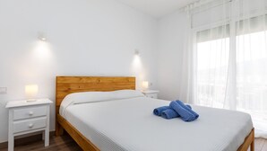 Un lit bien fait, avec une tête de lit en bois et une literie blanche, deux tables de chevet avec des lampes, et une grande fenêtre ornée de rideaux légers.