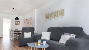 Un salon moderne avec un canapé gris, deux fauteuils, une petite table ronde sur laquelle sont posées des tasses et un gâteau, et un coin repas à l’arrière-plan.