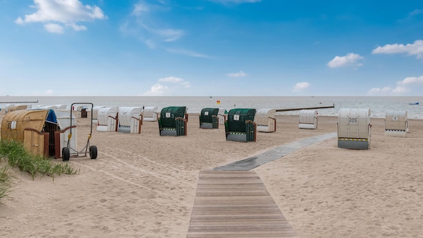 Ein Strand mit Liegestühlen, Sonnenschirmen, einer Holzpromenade und blauem Himmel.
