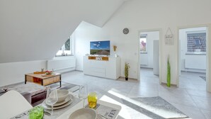 Ein geräumiges Wohnzimmer mit hohen Decken, einem Fernseher, einem Essbereich mit Tisch für zwei Personen und Blick ins Freie durch große Fenster.