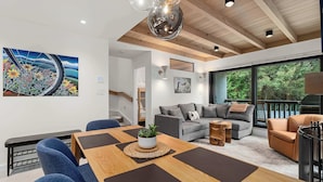 Un salon moderne avec une table à manger en bois, des chaises bleues, un canapé gris et une grande fenêtre donnant sur un paysage verdoyant.