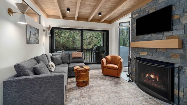 Un salon moderne avec une cheminée en pierre, un canapé gris, un fauteuil orange et une table d’appoint en bois.