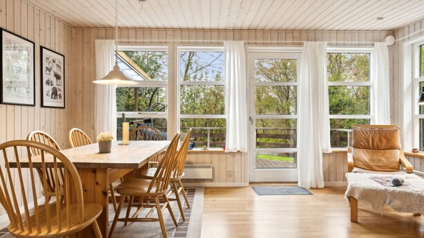 Une pièce chaleureuse avec une table à manger en bois, des chaises et un canapé. De grandes fenêtres sont ornées de rideaux, le sol est en bois et des tableaux encadrés décorent les murs.