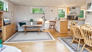 Un espace de vie et de salle à manger accueillant et ouvert, avec un plancher en bois, un canapé, une table à manger avec des chaises, un coin cuisine et de grandes fenêtres donnant sur un paysage verdoyant.