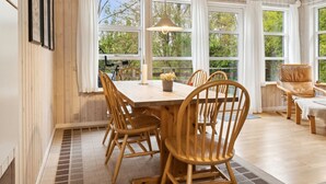 Un espace repas avec une table et des chaises en bois, de grandes fenêtres avec des rideaux et une vue sur la verdure à l’extérieur.