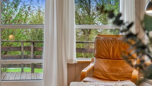 Une pièce dotée d’une grande fenêtre, d’une terrasse en bois à l’extérieur, d’un fauteuil confortable et d’une petite table sur laquelle se trouvent une couverture et du matériel de tricot.