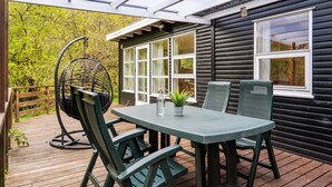 Une terrasse extérieure couverte, dotée d’une table et de chaises, d’un fauteuil suspendu et offrant une vue sur un chalet aux grandes fenêtres.