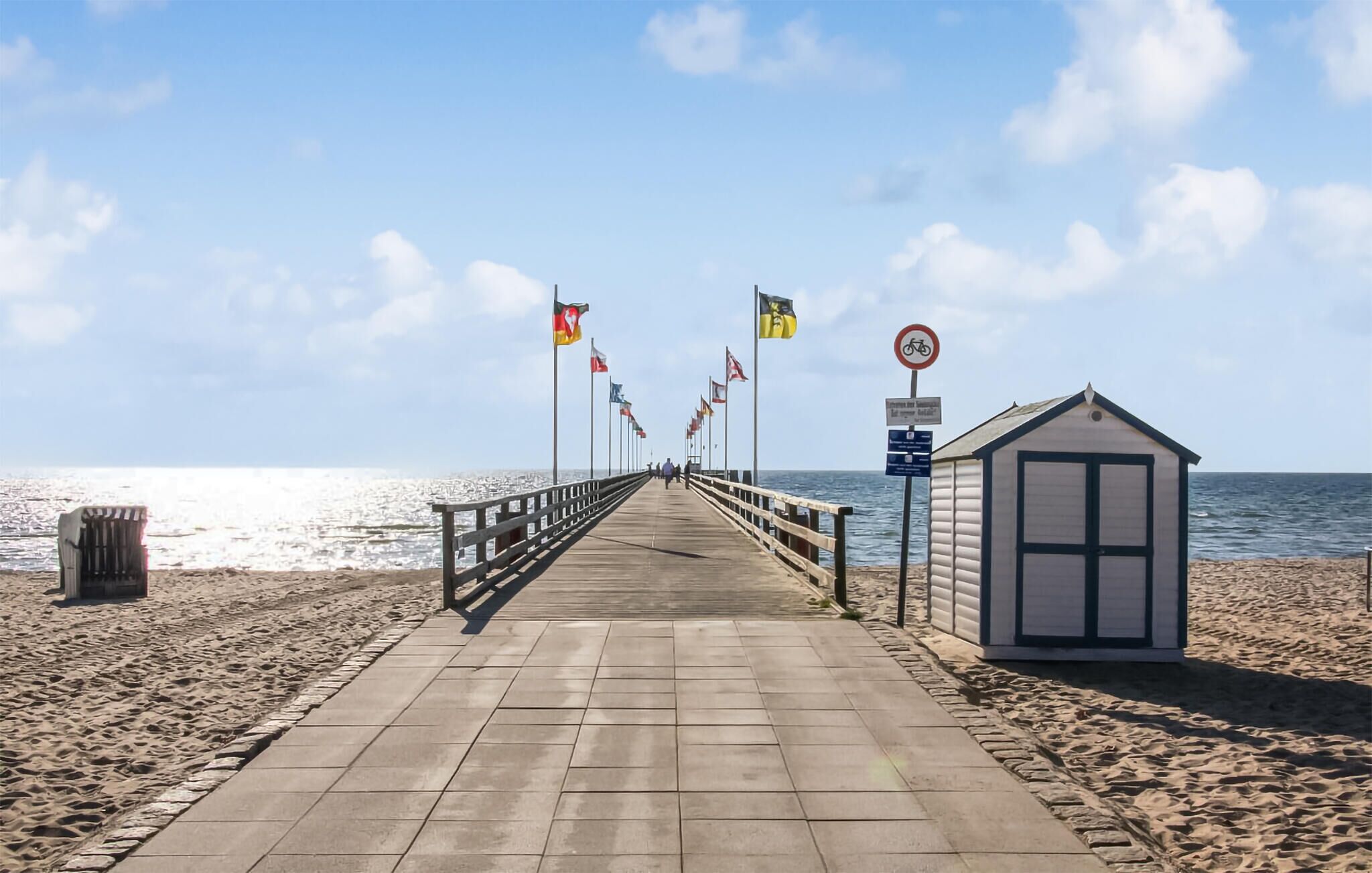 Une jetée en bois mène à une plage, et un petit bâtiment se trouve sur la droite. Des drapeaux sont visibles le long de la jetée.