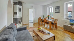 A spacious living area with a sofa, a wooden dining table set for four, and a kitchen with a decorative stove. - 3 bedroom cozy apartment in Großenbrode (Großenbrode)