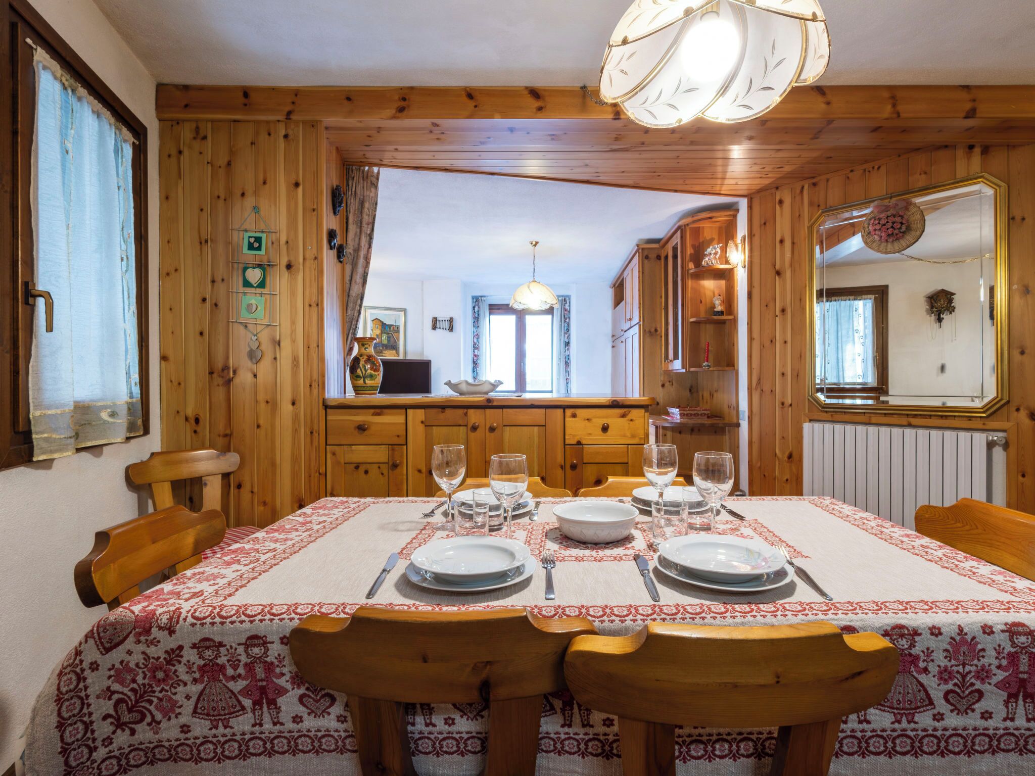 A dining area with a wooden table set for four, a chandelier, and a view into the kitchen.