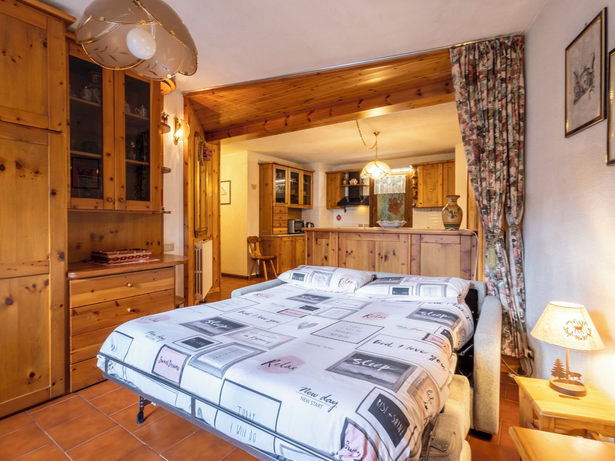 A cozy bedroom with a large bed, wooden cabinets, and a kitchen in the background.