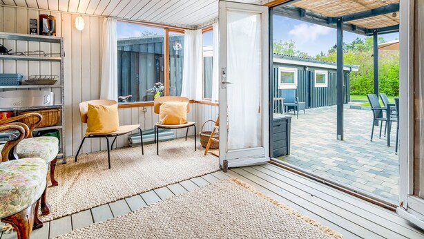 A room with a wooden ceiling, a sliding glass door leading outside, a small table with chairs, and a shelf with kitchenware.