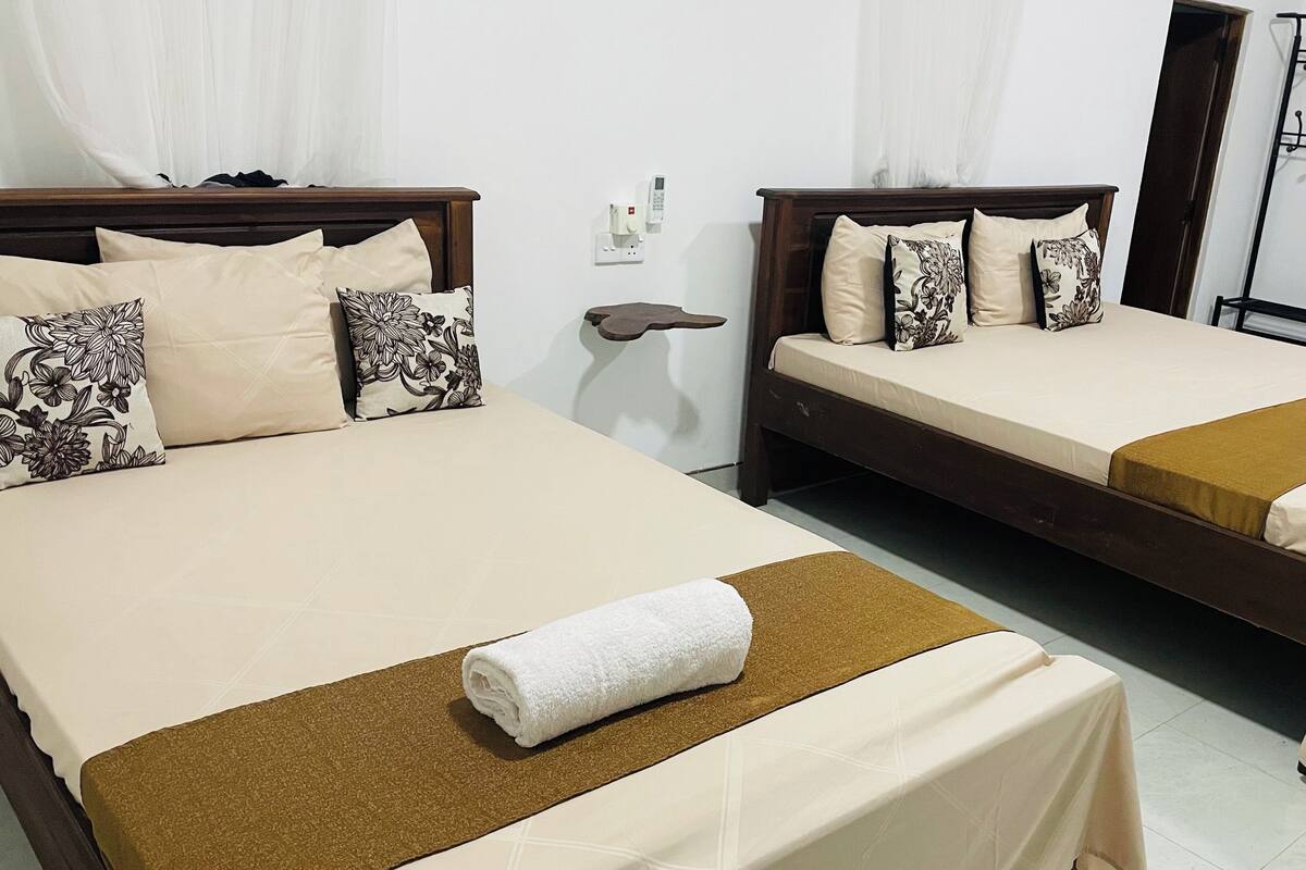 A hotel room with two beds, each with a brown headboard, beige bedding, and floral-patterned pillows. A white towel is placed on the bed in the foreground.