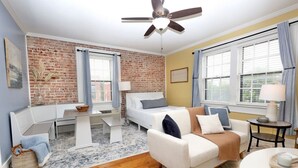 Una habitación acogedora con pared de ladrillo, cama, sofá blanco, una mesita y ventilador de techo.