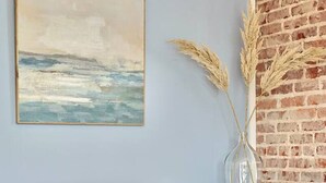 Una habitación con un banco blanco, un cuenco de madera sobre una mesa, un cuadro de paisaje marino y plantas secas en un jarrón.