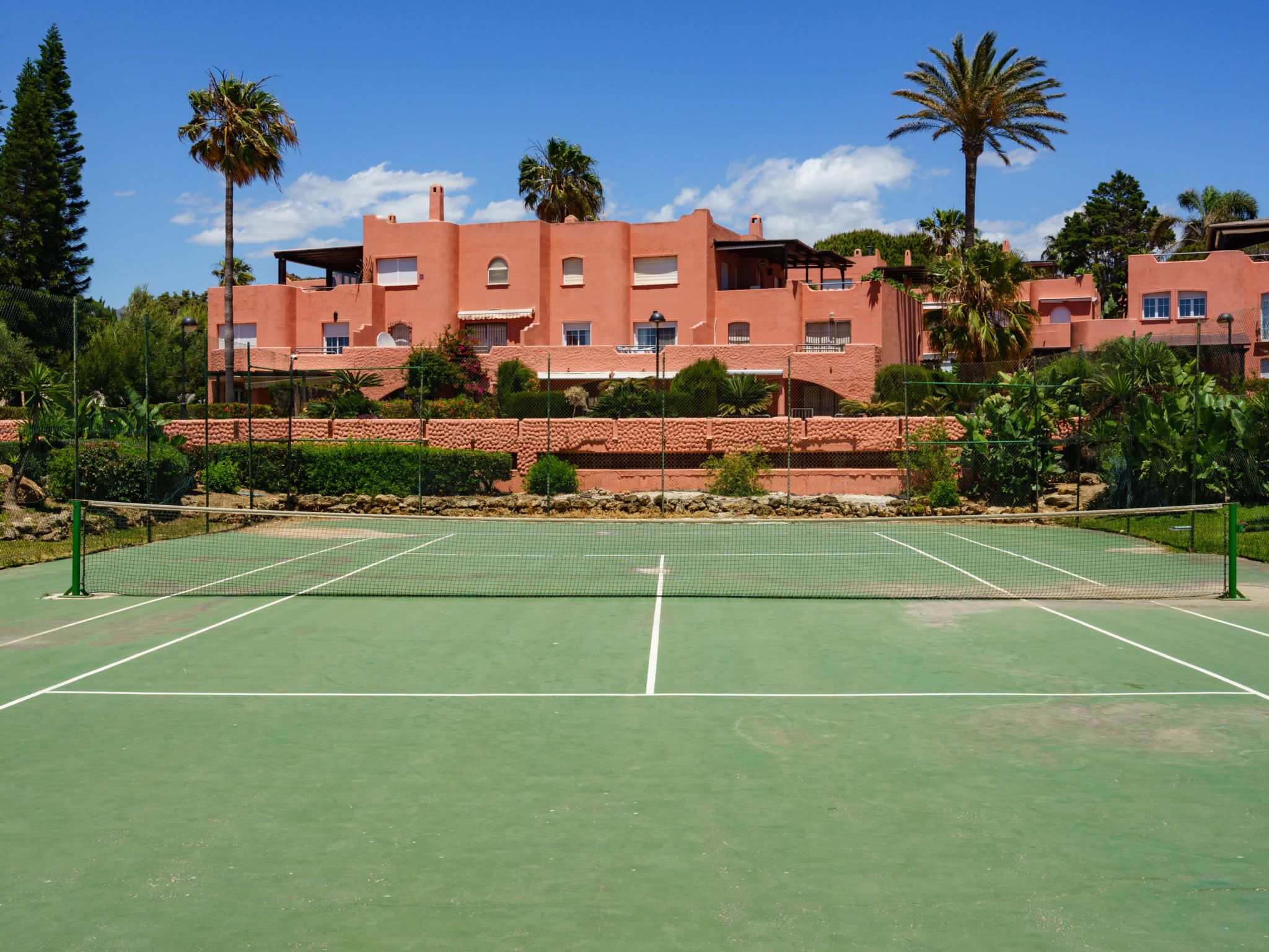 Una cancha de tenis con red, rodeada por una zona ajardinada con palmeras y un edificio al fondo con varias ventanas y balcones.