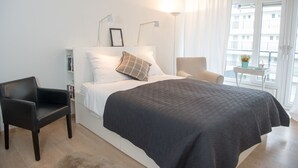 Ein Schlafzimmer mit einem Bett, einem Sessel, einem Nachttisch und einem Fenster mit Vorhängen.