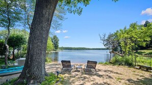 A lakeside setting with a large tree, two chairs, a small table, and a hammock.