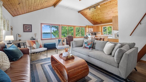 A spacious living area with a high ceiling, wooden beams, a large gray sofa, a wooden coffee table, and a view of a lake through large windows.