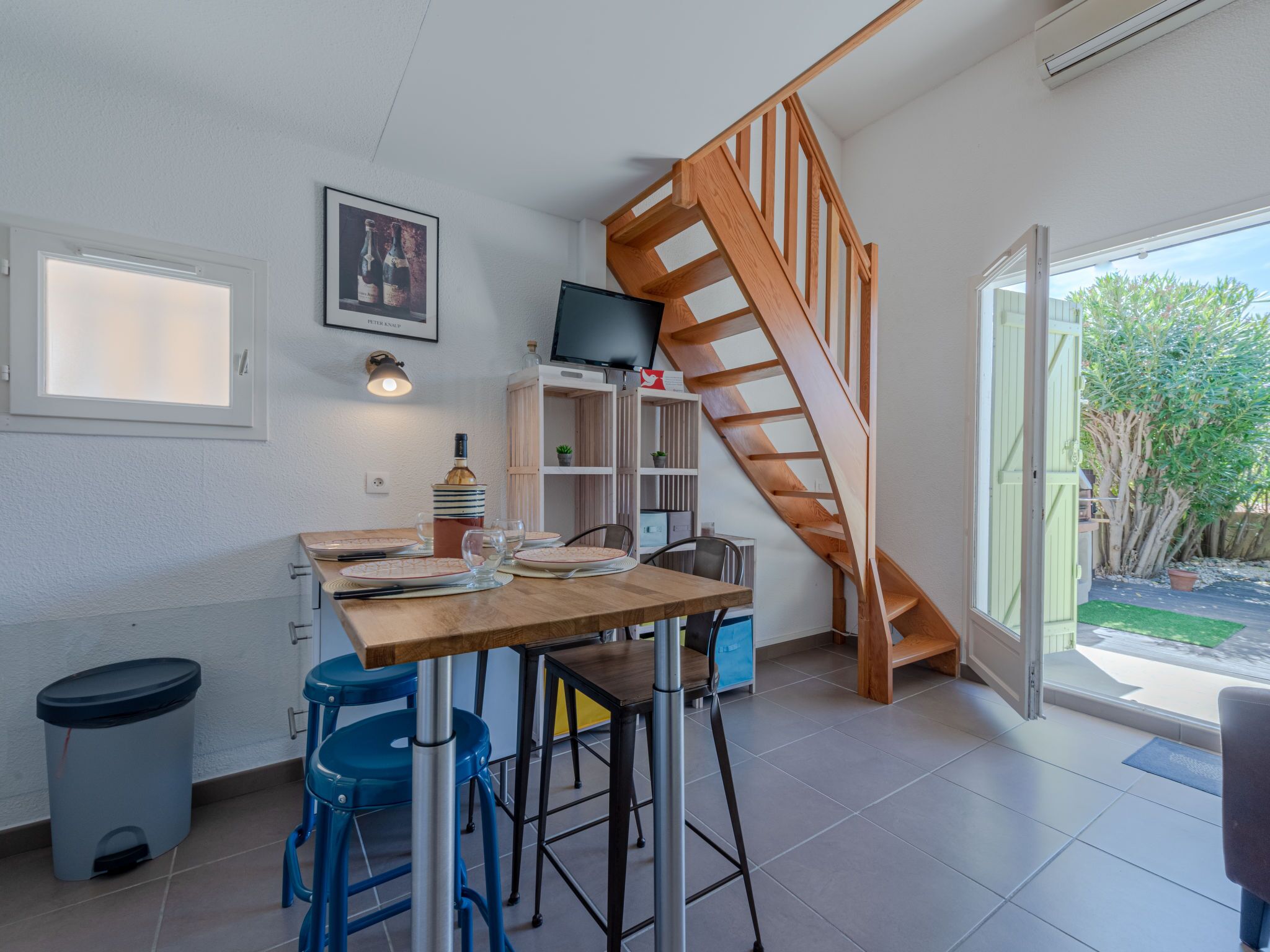 Un coin repas avec une table en bois, des tabourets de bar bleus, un téléviseur et un escalier en bois.