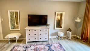 Un salon avec une commode blanche, un téléviseur à écran plat, un tapis à motifs et deux miroirs encadrés.