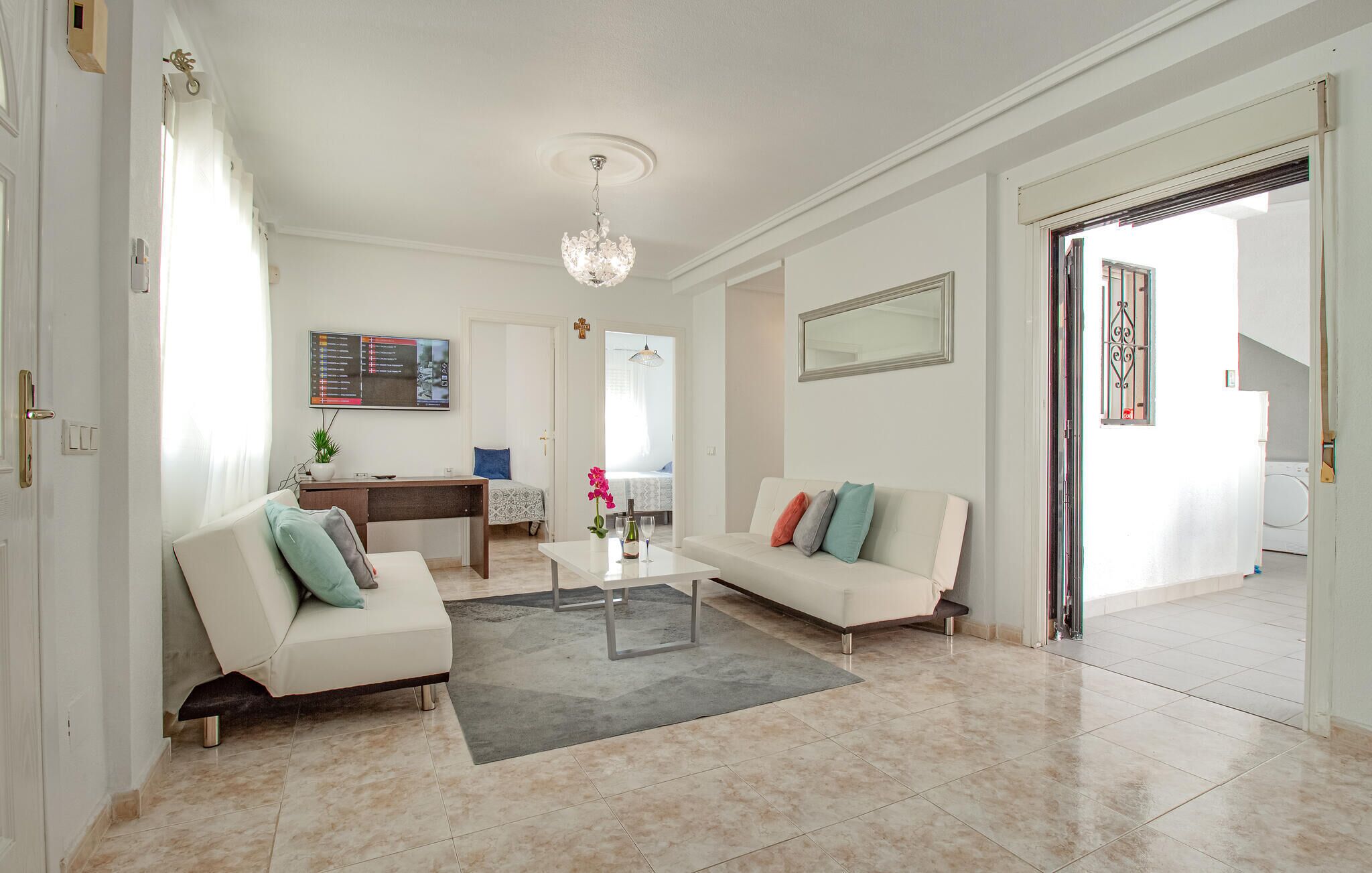 A spacious living room with a white sofa, a coffee table, and a chandelier. There is a dining area with a table and chairs, and a kitchen visible through an open door.