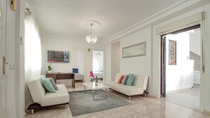 Amplia sala de estar con un sofá blanco, una mesa de centro y una araña. Se observa un comedor con mesa y sillas, y una cocina visible a través de una puerta abierta.