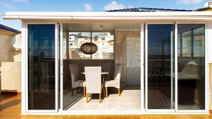Un moderno espacio exterior para comer con una mesa y sillas, una lámpara colgante y vistas de la ciudad a través de amplios ventanales correderas.