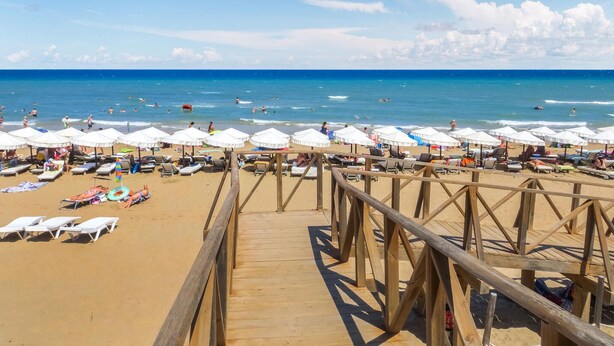 Pasarela de madera que conduce a una playa con sombrillas blancas y tumbonas.