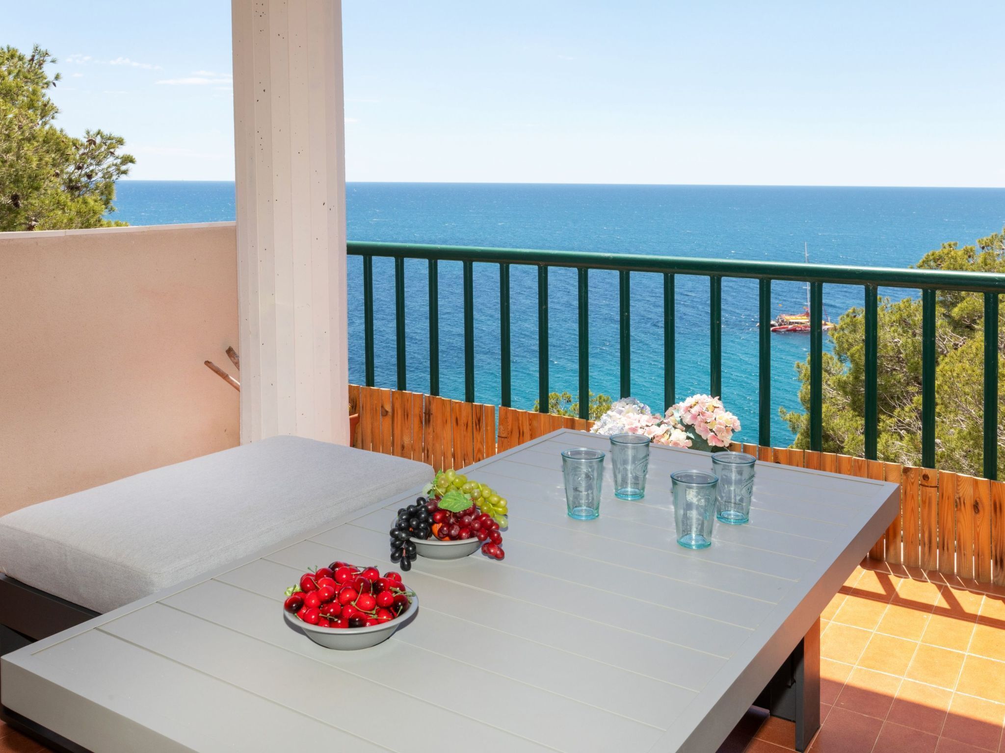A balcony with a table set for two, featuring a bowl of cherries and grapes, and a vase of flowers.